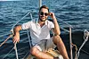 Smilende mann med skjegg og solbriller i hvit poloskjorte og lyse shorts sitter avslappet på dekk av en båt, med havet i bakgrunnen.