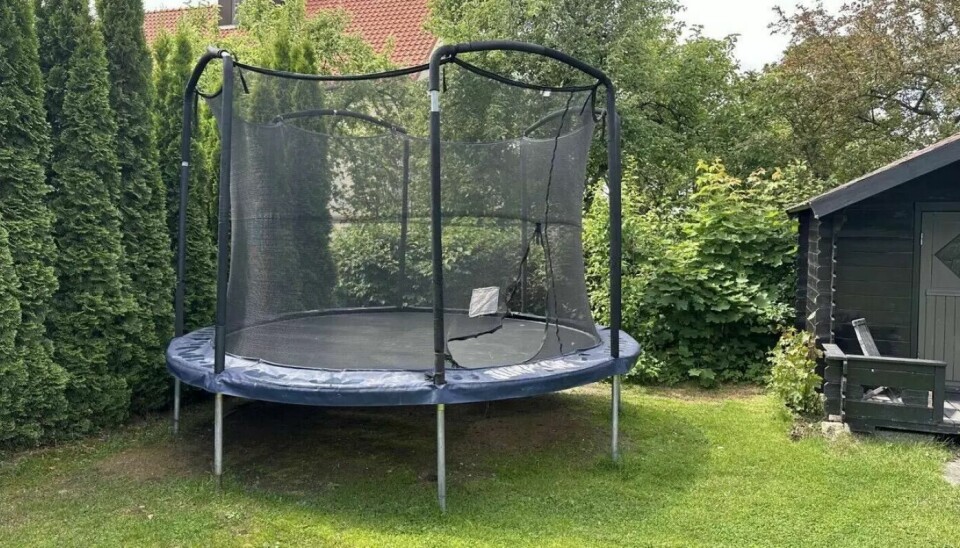 Trygg og morsom trampoline for hele familien. En god trampoline med sikkerhetsnett gir barna timevis med lek og bevegelse – og foreldre ro i sjelen. Trampoline med sikkerhetsnett plassert i hage med gressplen, omgitt av grønne busker og trær, ved siden av et lite mørkt hagehus.