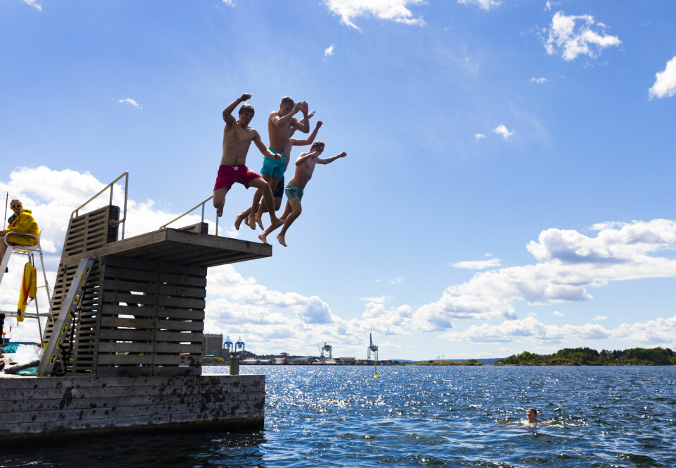 Sol, skyer og bading på Sørenga i Oslo, et av stedene som kan være ekstra utsatt for forurensning i sommer. Fire gutter hopper samtidig ut fra et stupetårn i sjøen på en solrik dag. En badevakt i gult sitter til venstre, og én person er synlig i vannet. Det er blå himmel og glitrende sjø i bakgrunnen. Sommerglede og stup fra bryggekanten – men visste du at vannet kan inneholde farlige bakterier etter styrtregn? Eksperter anbefaler å vente med bading etter kraftige regnskyll, spesielt i bynære områder.