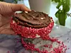 En hånd holder en sandwichis laget med sjokoladecookies og vaniljeis, rullet i rosa jordbærstrø og servert på en marmorplate. Enkel og dekorativ dessert til varme dager.