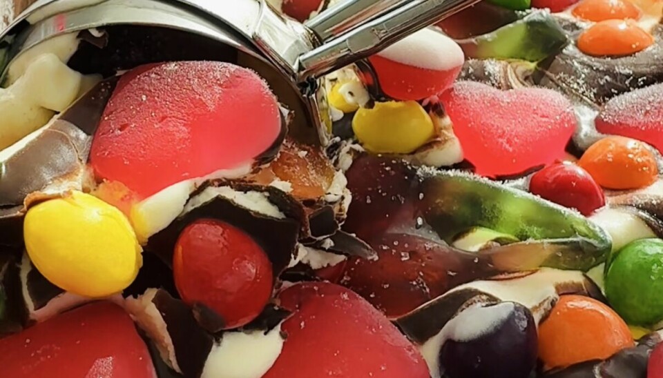 Denne smågodt-isen lager du helt selv – og det beste er at du kan bruke akkurat det godteriet du liker aller best. Nærbilde av hjemmelaget iskrem laget med kondensert melk og kremfløte, toppet med sjokoladesaus som stivner og store mengder smågodt som geléhjerter, sjokoladeperler og fargerike dragéer. En isskje løfter en porsjon ut av formen.