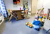Et lite barn med blondt hår, sett bakfra, sitter på gulvet i et rom fylt med leker. Barnet leker med et gult lekekjøretøy og et fargerikt lekesett. Rundt barnet står det flere trestoler og bord, og rommet har et barnehagepreg med lyse vegger og barnevennlig innredning.