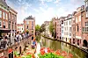 Bildet viser en idyllisk kanal i gamlebyen i Utrecht, Nederland, omgitt av sjarmerende murbygninger, utekafeer og blomster i forgrunnen.