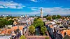 Bildet viser en panoramautsikt over Utrecht, Nederland, med den ikoniske Domkirketårnet (Domtoren) som rager opp over byens lave bygninger med oransje tak. I forgrunnen slynger en grønn kanal seg gjennom bykjernen, omgitt av trær og sjarmerende historiske hus.