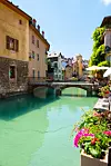 Et idyllisk kanalparti i Annecy, Frankrike, med klare turkise vann omgitt av pastellfargede bygninger i gult, oransje og grønt. Til høyre pryder blomsterkasser med fargerike blomster kanalkanten, mens en liten steinbro med to buer binder sammen sidene. I bakgrunnen strekker kanalen seg videre mellom sjarmerende gamle hus, med en klar blå himmel over som gir en varm og livlig atmosfære.