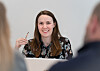 Her ser vi et portrettbilde av en kvinne som smiler vennlig. Hun sitter ved et bord med en bærbar datamaskin foran seg, og holder et par briller i den ene hånden. Hun har langt brunt hår og har på seg en mørk, blomstermønstret bluse. Bildet er tatt i et lyst rom med en nøytral bakgrunn, noe som gir et profesjonelt og ryddig preg.

Fokuset er på henne, mens to personer i forgrunnen er uskarpe, noe som gir inntrykk av at hun er i en samtale eller et møte. Stemningen fremstår positiv, profesjonell og inkluderende.