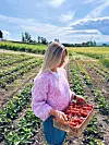Modell iført «Midtsommer»-genseren fra By Wergeland – en håndstrikket genser i melert rosa garn. Bildet er tatt på en jordbæråker i norsk sommersol, og fanger det landlige, fargerike uttrykket i både plagget og omgivelsene.