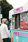 En mann bestiller iskrem fra en fargerik isbil merket 'Akhi’s Ice Cream Van'. Bilen er dekorert med menyer og illustrasjoner. Nøkkelord: Akhi’s Ice Cream, street food London.