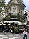 Fasade til Café de Flore i Paris med inngang og rundtliggende bygninger. Dette bildet viser fasaden til den berømte kaféen Café de Flore i Paris. Kaféens navn vises tydelig på skilt og markiser. Fasaden har dekorative balkonger, vinduer med smijernsrekkverk og grønt planteinnslag over kaféområdet. Nærbilde av bygninger rundt og gatebelysning samt mennesker som passerer bidrar til gateatmosfære i by.