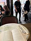 Bord på Café de Flore med bok og bestikk i forgrunnen, uteservering i Paris. På bildet ser man et rundt kafébord med dekorert bordplate hvor teksten “Café de Flore” er synlig. På bordet ligger en åpen bok med sider vendt mot kamera, og til høyre ligger kniv og gaffel lagt på en serviett. I bakgrunnen skimtes uteservering med stoler, bord og mennesker i Paris, litt uskarpt. Bildet gir assosiasjoner til rolig kaféstund med lesing og atmosfære i Paris.