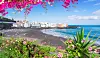 Utsikt over en strand med svart sand, turkis hav og fargerike hus langs kysten på Kanariøyene, omgitt av blomstrende bougainvillea