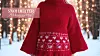 En kvinnelig modell iført en rød strikket ponchogenser med hvite hjerte- og snøkrystallmønstre, fotografert foran et vinterlig bakteppe med snødekte trær og varme lysdekorasjoner. Genseren har vide ermer og høy hals, og passer perfekt til jul og kalde vinterdager.