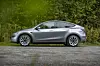 Et sidebilde av en grå Tesla Model Y parkert utendørs på grusvei, omgitt av grønn vegetasjon. Bildet viser bilens moderne design med strømlinjeformet karosseri og minimalistiske detaljer.