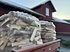 Flere store sekker med ved stablet på tilhenger foran en rød gårdsbygning. Bildet illustrerer produksjon og levering av fyringsved. Nøkkelord: vedlevering, fyringsved.