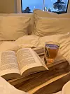 Bilde av en bok og en kopp te på et trebrett i en seng. Sengen har hvitt sengetøy på, og de tre elementene, lesing, noe varmt å drikke og myke tekstiler, er en del av det som kalles «cozymaxxing»