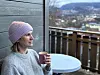 Bilde av en kvinne i en grå stikkegenser og med en lilla strikkelue på hodet, som sitter på en terrasse og titter utover et område utenfor leiligheten ved Bergen på Vestlandet. Hun holder en kopp i handen og bak henne ser vi fjell og hus som kryper oppover fjellsiden.