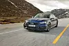 Blå Audi RS5 i fart på øde fjellvei med fjell i bakgrunnen.