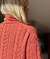 Nærbilde bakfra av person i oransje flettestrikket genser med høy hals.