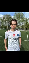 Fotballspiller i hvit drakt står på kunstgressbane med trær i bakgrunnen