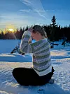 Person med stripet, hjemmestrikket genser sitter i snøen ved skog i solnedgang.