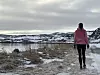 Person i rosa jakke går på vinterlig sti langs kysten med fjell og sjø i bakgrunnen.