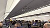 Fullsatt flykabine med passasjerer sittende i seteradene under bagasjehyller. Reise.