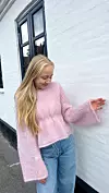 Ung kvinne i rosa, håndstrikket genser og jeans står ved en hvit murvegg.