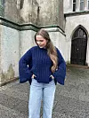 Person utendørs iført mørkeblå, oversized flettestrikket genser og lyse jeans foran murbygning.