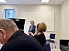 Eldre kvinne står foran kursdeltakere i lyst møterom med stoler og whiteboard.