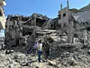Flere personer går gjennom ruiner av sammenraste bygninger i et ødelagt byområde.