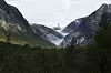 Nigardsbreen som lyseblå isarm mellom bratte fjell over en grønn dal med skog.