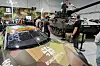 Bundeswehr-stand med kampvogn og kamuflert racingbil omgitt av publikummere innendørs på messe.