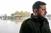 Portrett av Volodymyr Zelenskyj i grønn jakke på en flyplass med fly i bakgrunnen.