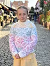 Barn iført flerfarget, mønsterstrikket genser står i en solfylt brosteinsgate.