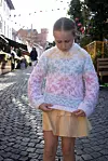 Jente i pastellfarget strikkegenser står i en solfylt brosteinsgate og ser ned på genseren.