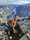 Person lager mat på primus på fjelltopp med dyp fjord og fjell i bakgrunnen. Influencer Johan Haugsrud lager mat på primus ved fjellkanten Bakkanossi med spektakulær utsikt over den norsk fjorddalen, Nærøyfjorden.