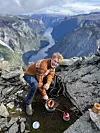 Person lager mat på primus på fjelltopp med dyp fjord og fjell i bakgrunnen. Influencer Johan Haugsrud lager mat på primus ved fjellkanten Bakkanossi med spektakulær utsikt over den norsk fjorddalen, Nærøyfjorden.
