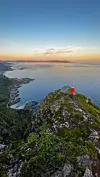 Person i rødt ved telt på smal fjellryggen Børtinden i Andøya, med utsikt over hav og kystlinje i solnedgang. En turgåer med telt slår leir på en smal fjellrygg med utsikt over kysten, i jakt på nye eventyr utenfor de mest kjente turiststiene.