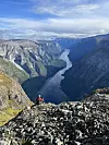 Person sitter på steinete fjellkant på Bakkanossi ved Nærfjord. med utsikt over dyp fjord mellom høye fjell. En turgåer tar pause på fjellkanten med vid utsikt over en dyp norsk fjord, slik eventyreren Johan anbefaler i sine turforslag.