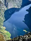 Utsikt fra høyde ned mot dyp blå fjord med båt og bratte, grønne fjellsider. Turentusiast utforsker norsk natur på fjellturen Bakkanossi, med utsikt over en dyp Nærøyfjoren i Aurland kommune i Vestland fylke. I bakgrunnen ser man en båt som skjærer gjennom blankt vann mellom bratte fjellsider.