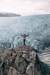 Person står på bergknaus foran stor isbre med armene utstrakt. En enslig turgåer med rødt telt camper ved isbreen Svelgabreen i Kvinnherad i Vestland fylke, og elv, på jakt etter nye norske naturopplevelser utenfor de mest kjente rutene.