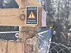 Nærbilde av trestolpe med advarselsskilt ved inngang til hengebru i skogen.