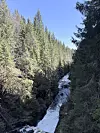 Trang elv som fosser gjennom et skogkledd juv under blå himmel, over hengebrua ved Bjørnsjøhelvete. Dramatisk elvejuv omgitt av tett granskog viser hvorfor Oslomarka frister til korte eventyrturer for barn og voksne.