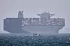 Stort containerskip ved anker i disig vær mens en liten motorbåt passerer foran i Hormuzstredet.