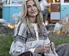 Person i mønstret Setesdal-jakke sitter i gresset og strikker foran en campingbil.