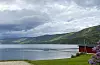 Landskap ved Holsfjorden med naust, brygge og grønne åser under skyet himmel.