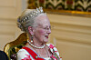 Dronning med tiara og perlesmykker sitter på en gyllen stol i et pyntet salongrom.