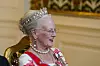 Dronning med tiara og perlesmykker sitter på en gyllen stol i et pyntet salongrom.