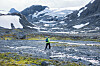 Person krysser bekk i steinete høyfjell med snø og bre i bakgrunnen.
