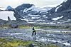 Person krysser bekk i steinete høyfjell med snø og bre i bakgrunnen.